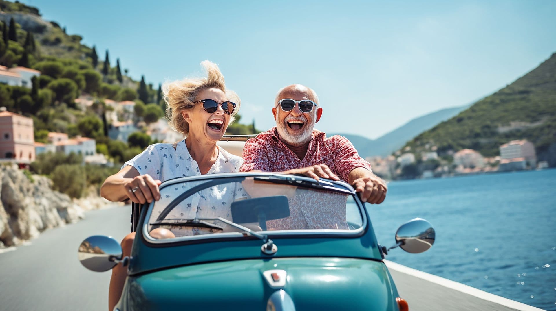 Un couple âgé rit en conduisant une voiture ancienne sur une route côtière ensoleillée.