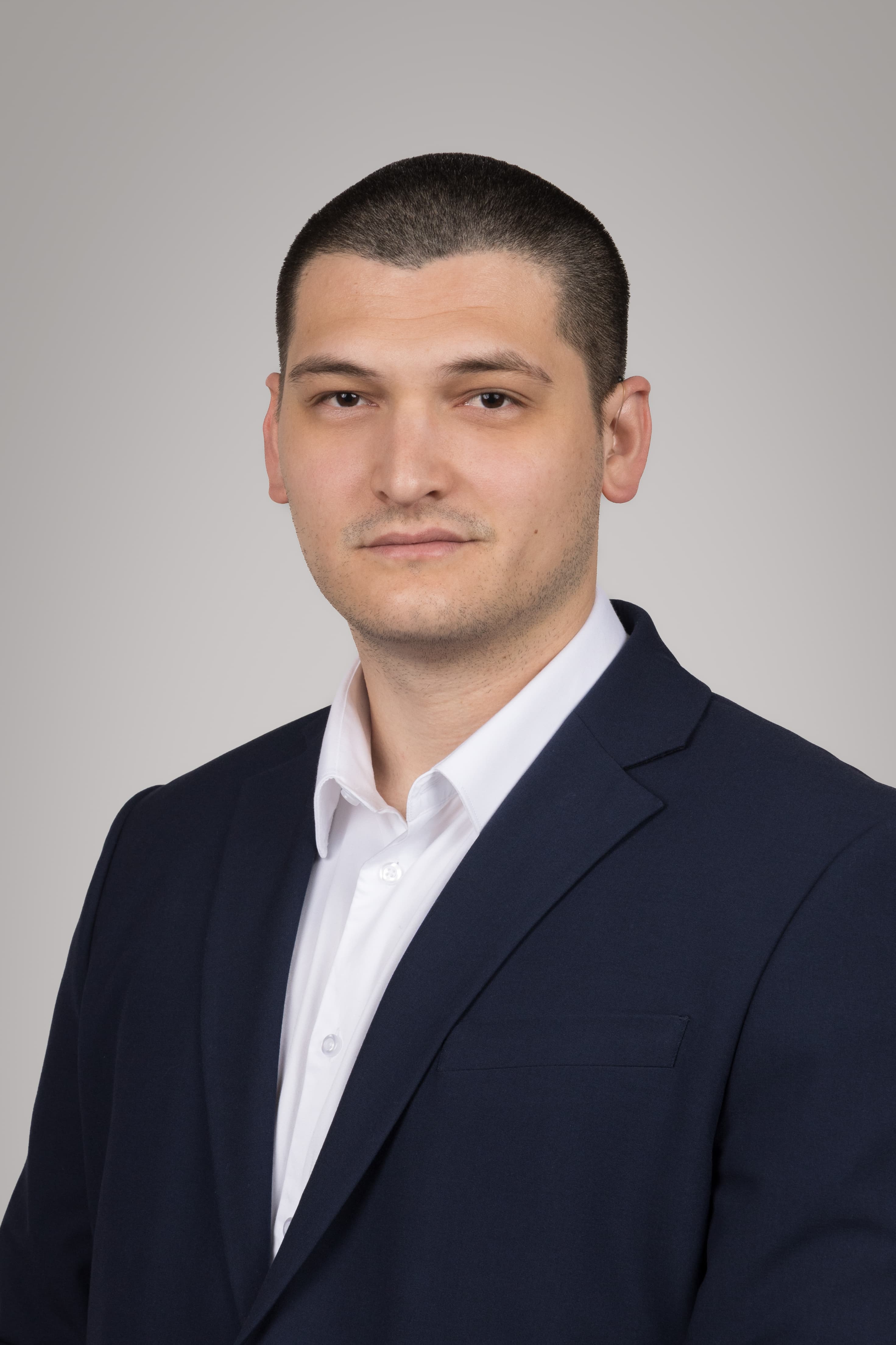 Homme aux cheveux courts en chemise blanche et veste bleu marine sur fond gris.